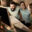Three coworkers celebrate at a desk, smiling and cheering in front of a large computer monitor. They appear happy and excited, working together in a modern office with exposed brick walls.